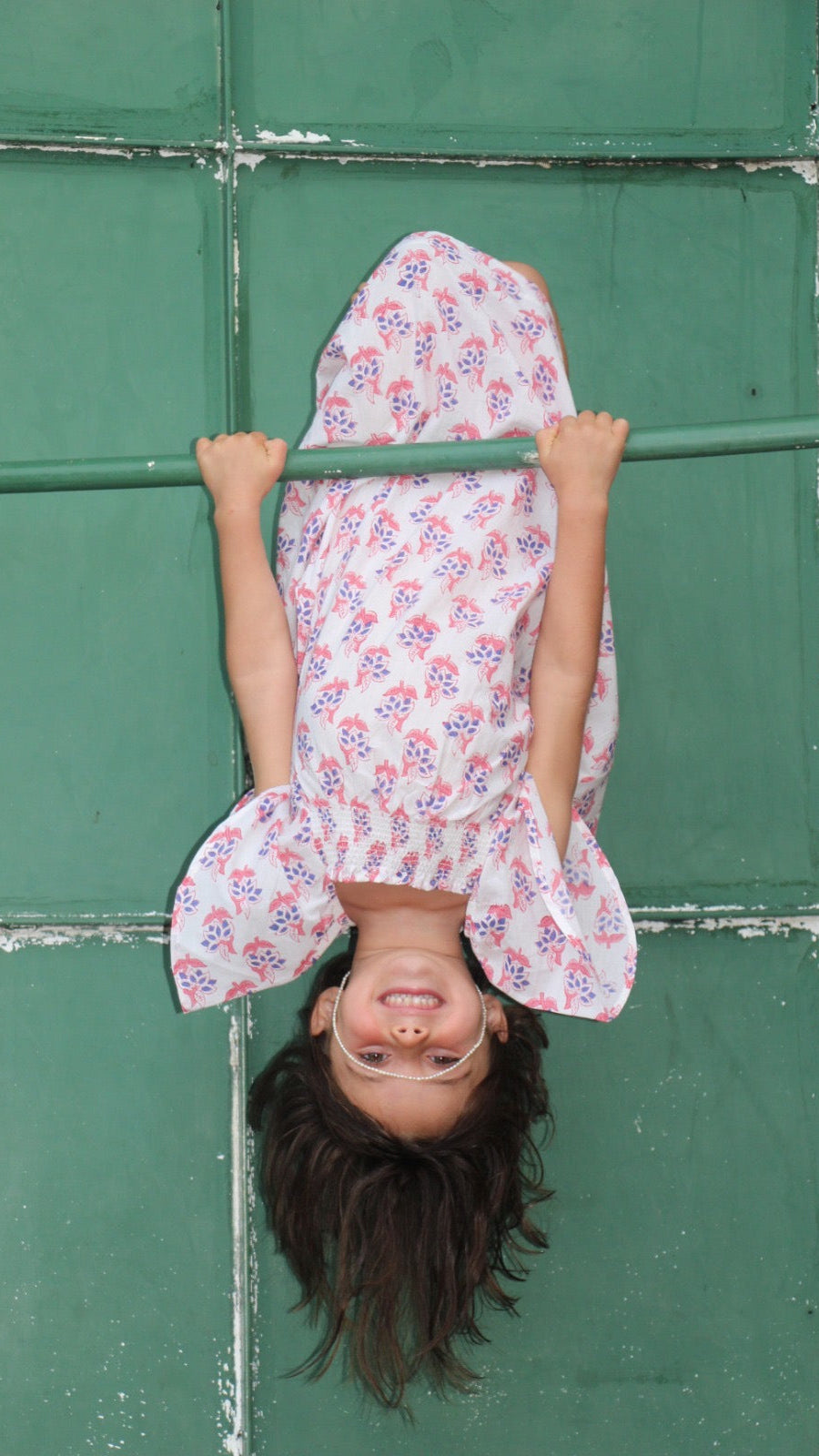 Child in a floral dress hanging upside down against a green wall